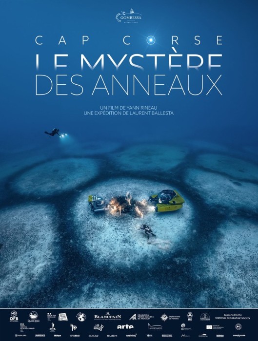 Affiche de film documentaire sous-marin, dominée par des tons bleu profond. En haut, le titre « CAP CORSE – LE MYSTÈRE DES ANNEAUX DES ANNÉAUX » est inscrit en grandes lettres blanches. Le sous-titre indique : « Un film de Yann Rineau » et « Une expédition de Laurent Ballesta ». Au centre, un plongeur équipé évolue au-dessus d’un vaste paysage sous-marin circulaire. Au premier plan, plusieurs plongeurs et équipements scientifiques entourent une zone claire sur le fond marin, avec notamment un petit engin jaune. De nombreux logos de partenaires et organismes figurent en bas de l’affiche.